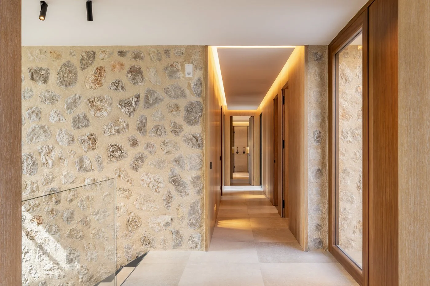 Pasillo moderno con paredes de piedra y madera, iluminación cálida y ventanas grandes.
