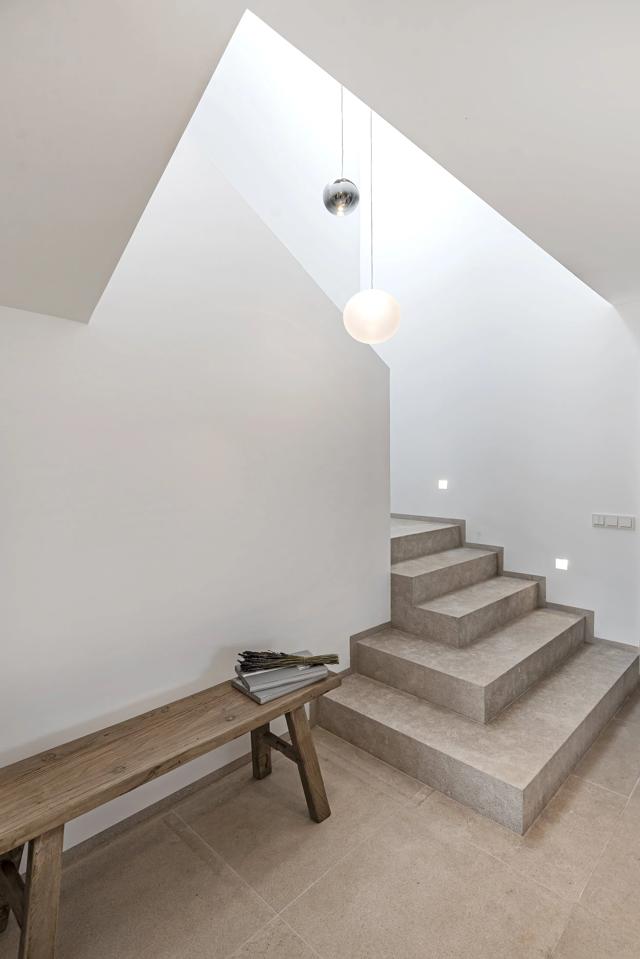 Interior de una escalera moderna con paredes blancas, iluminación natural desde una ventana en el techo, escalones de piedra y una banca de madera con algunos papeles encima.