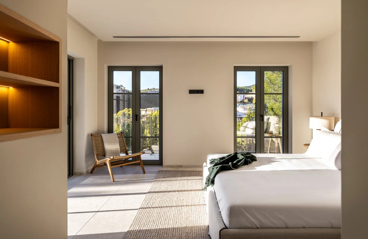 Habitación con cama blanca, lámpara, sillas y ventanas con vista a la ciudad y naturaleza.