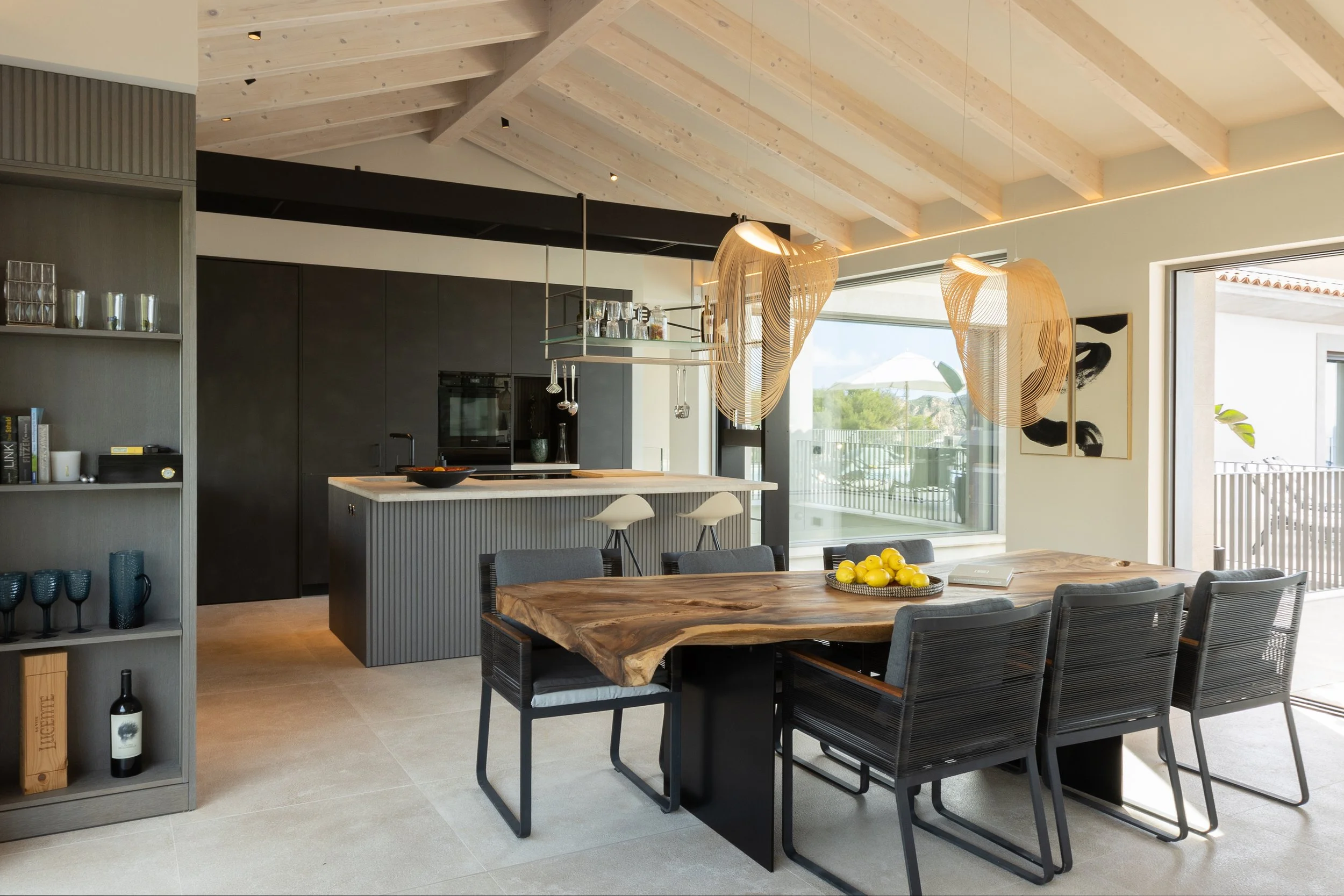Interior de una cocina moderna con paredes blancas, techo de madera y una isla central gris, decorada con lámparas de diseño ondulado en tonos madera y una mesa grande de madera con sillas negras. Grandes ventanas permiten la entrada de luz natural.