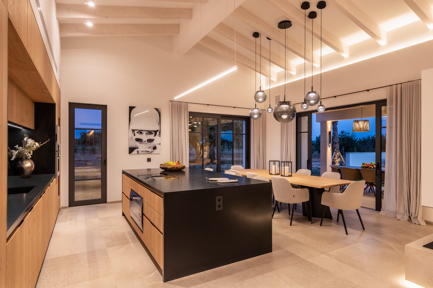 Cocina moderna con isla negra, comedor con sillas blancas, puertas corredizas de vidrio hacia el exterior, decoración minimalista y lámparas colgantes de metal.