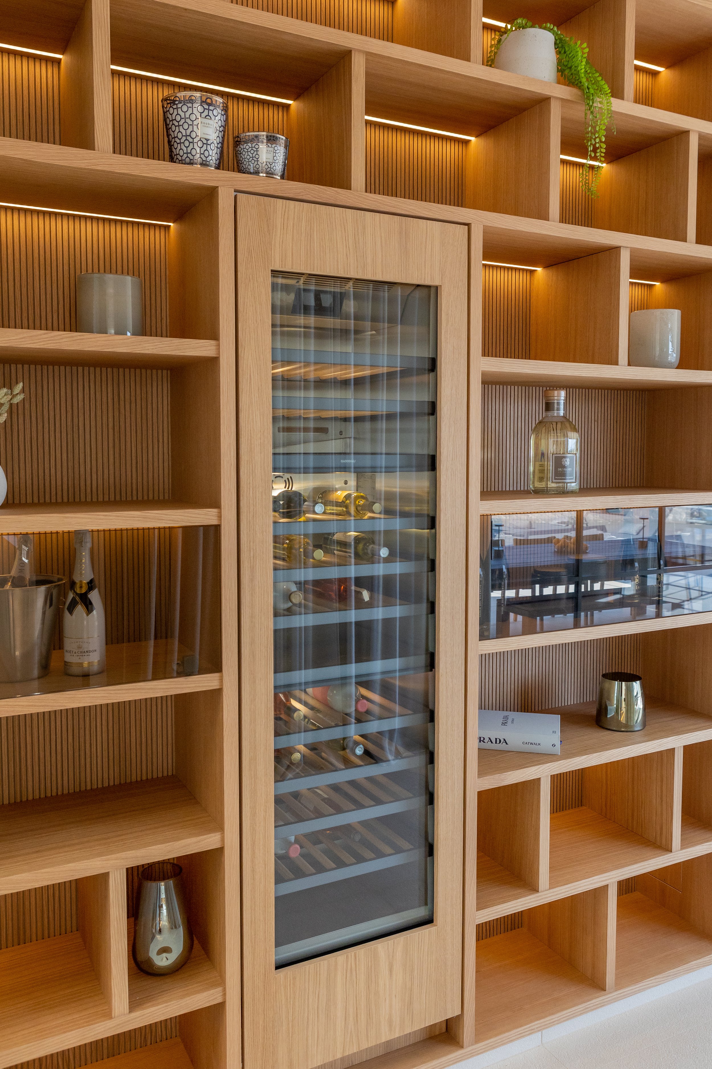 Estantería de madera con botellas de vino, vasos y decoraciones, incluyendo plantas y libros.
