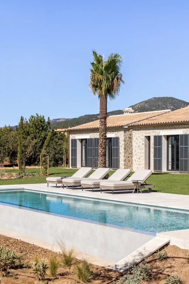 Casa de piedra con ventanas de persianas negras, piscina con tumbonas, palmera, y montañas al fondo.