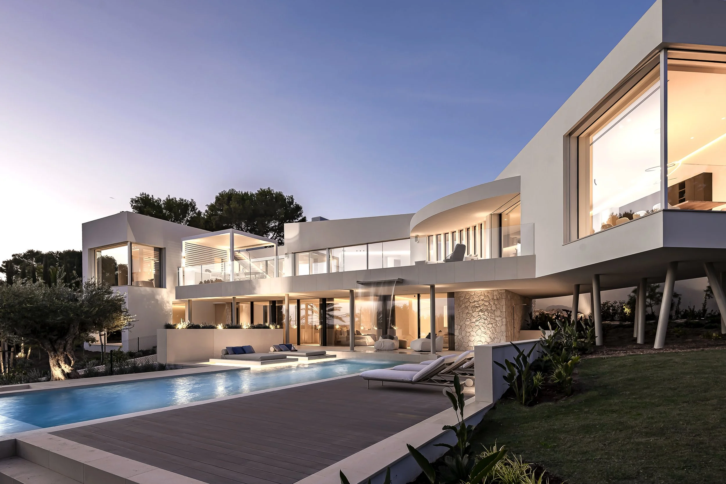 Casa moderna con muchas ventanas grandes, piscina y áreas de descanso exterior al atardecer.