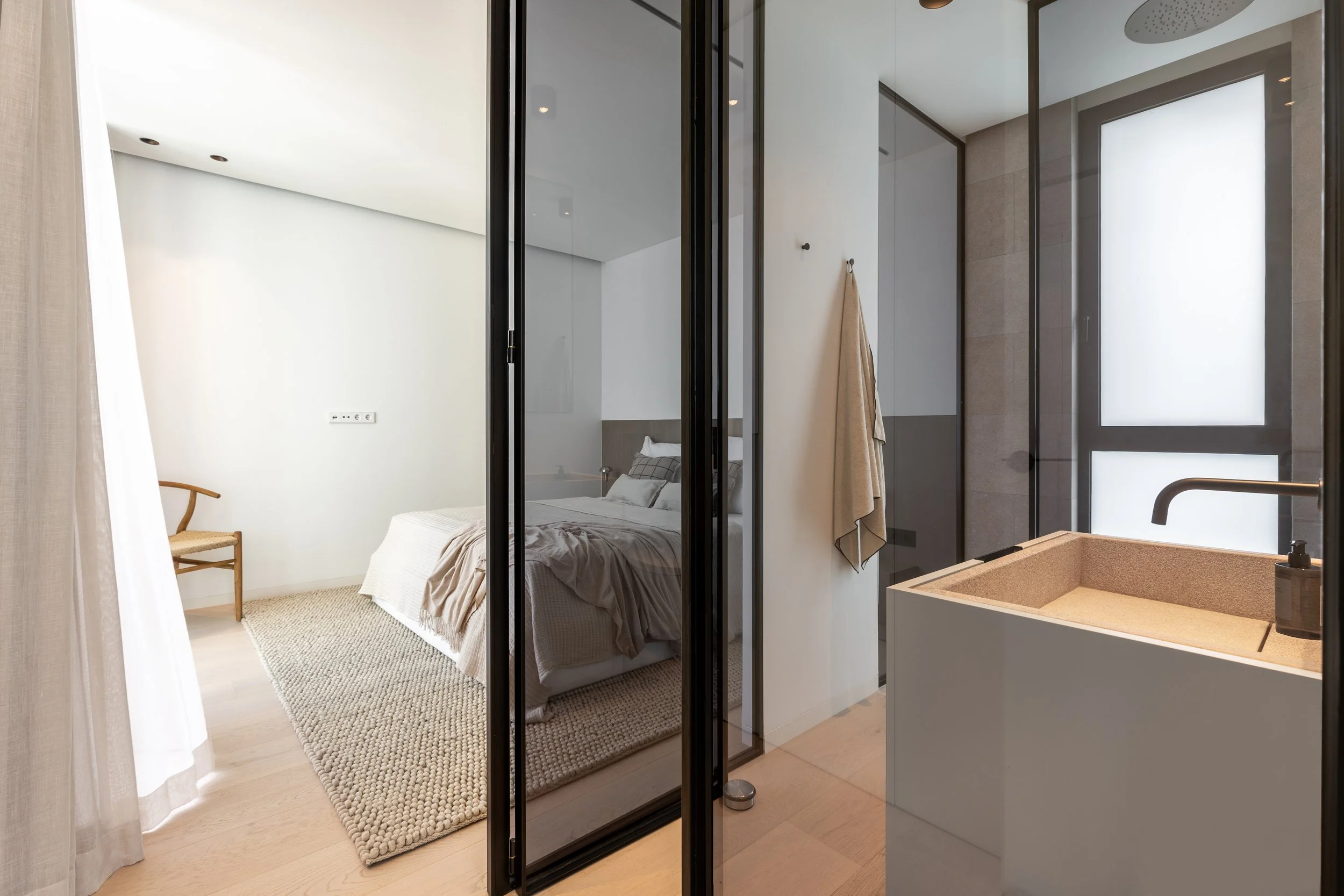 Vista de una habitación moderna desde la cocina, con una cama, una silla de madera, un tapete beige, paredes blancas, un armario con puertas de espejo y una ducha con puerta de vidrio.