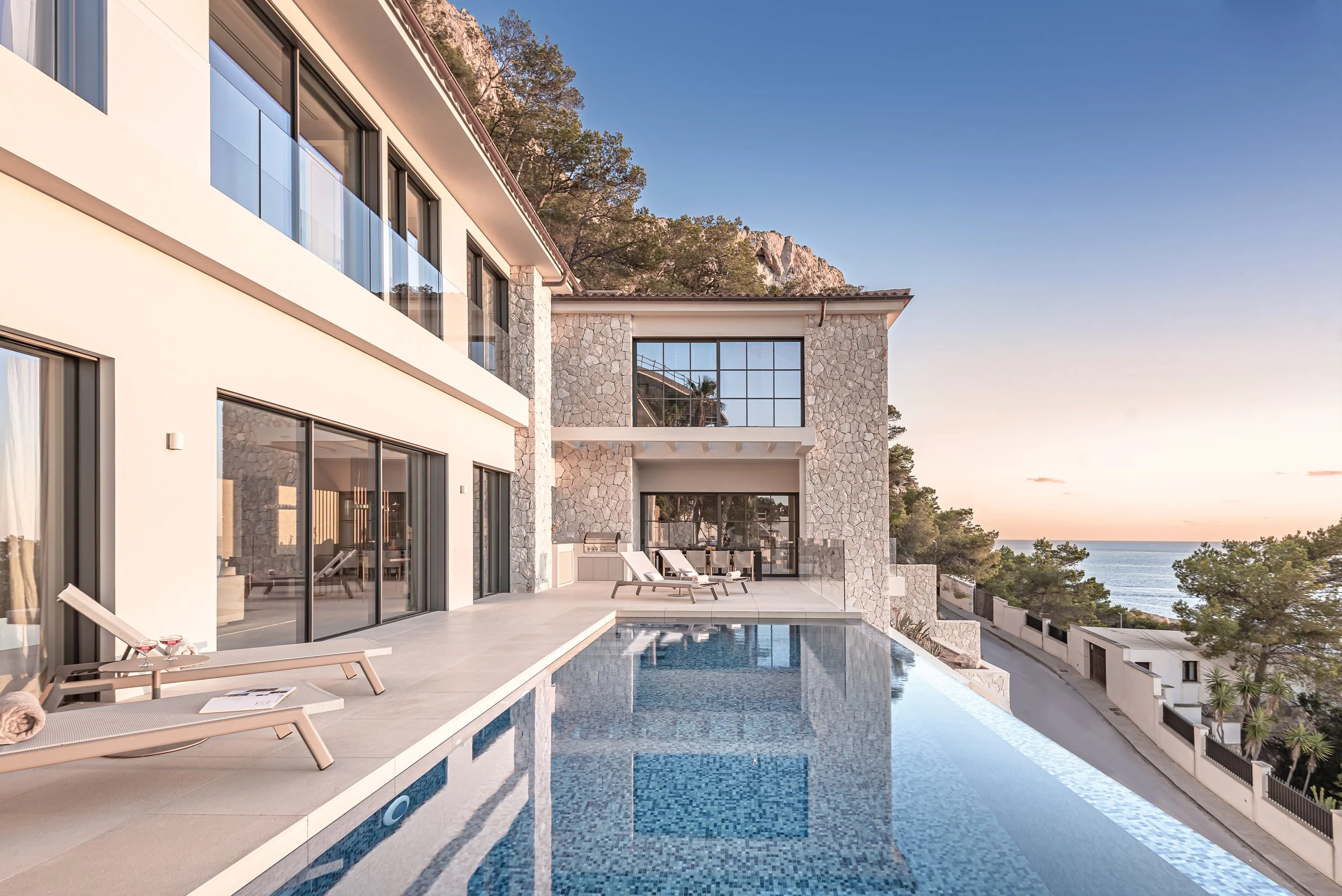 Vista de una piscina moderna frente a una casa con paredes de piedra y ventanas de vidrio, con una vista del mar y montañas al fondo, durante el atardecer.