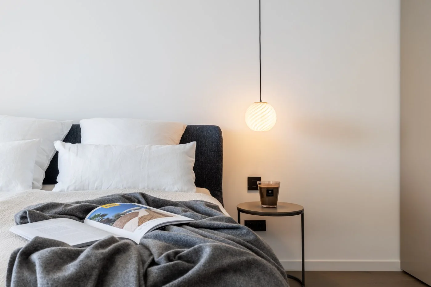 Cama con almohadas blancas y una revista abierta sobre una manta gris, lado de una lámpara colgante y una taza en una mesita redonda