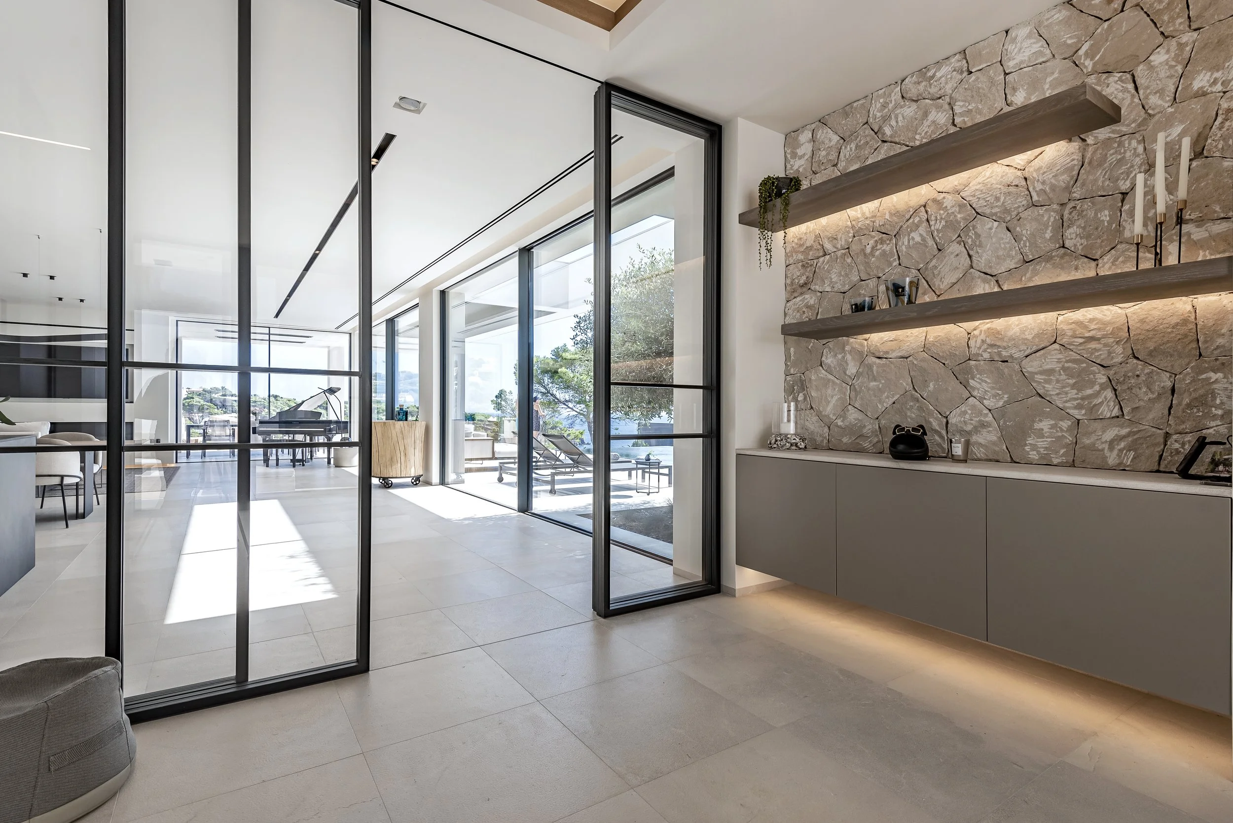 Interior de una sala con paredes de piedra y estanterías de madera, dividida por puertas de cristal y marco negro, permite vista hacia un espacio con un piano y vistas al exterior con árboles y cielo.