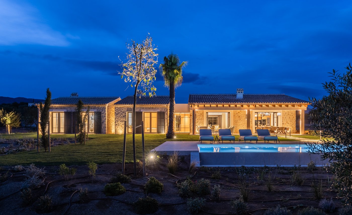 Casa iluminada con piscina y área de descanso en la noche, rodeada de árboles y jardín.