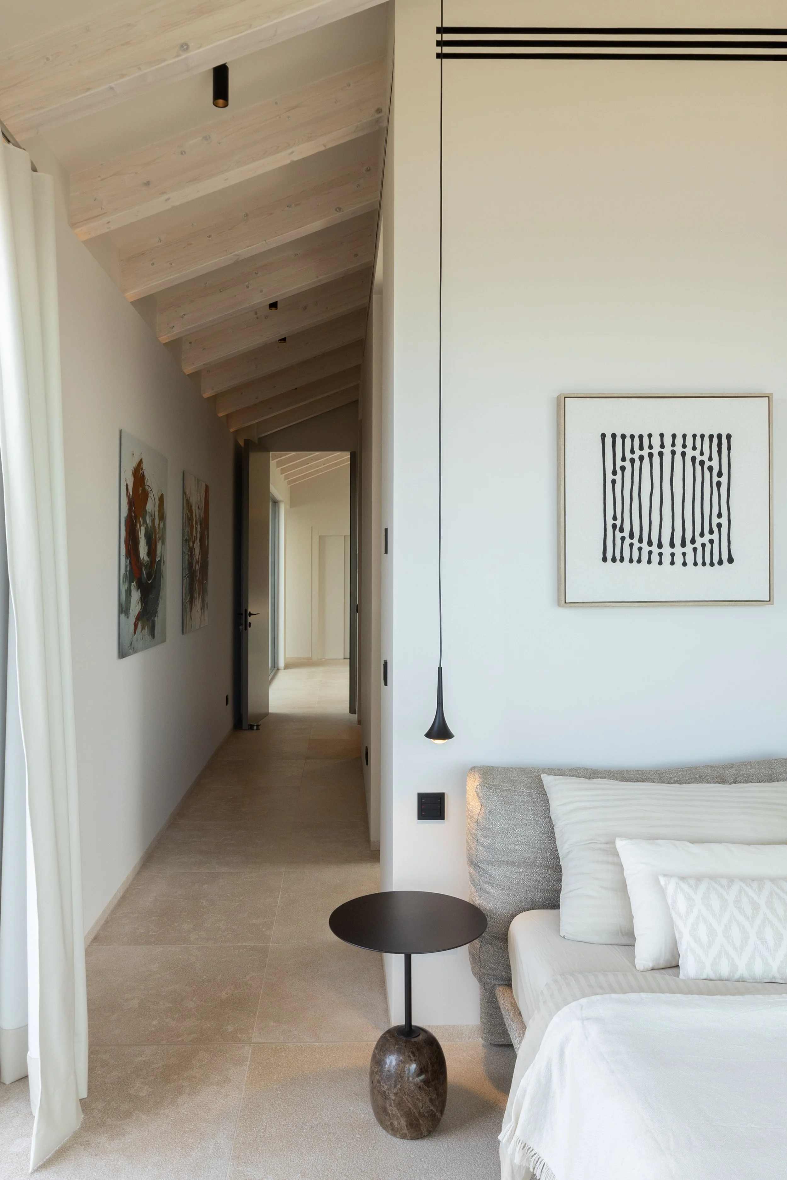 Interior de una habitación moderna con paredes blancas, una cama con ropa blanca, una lámpara colgante negra, y cuadros abstractos en la pared. Se ve un pasillo que lleva a otra zona de la casa.