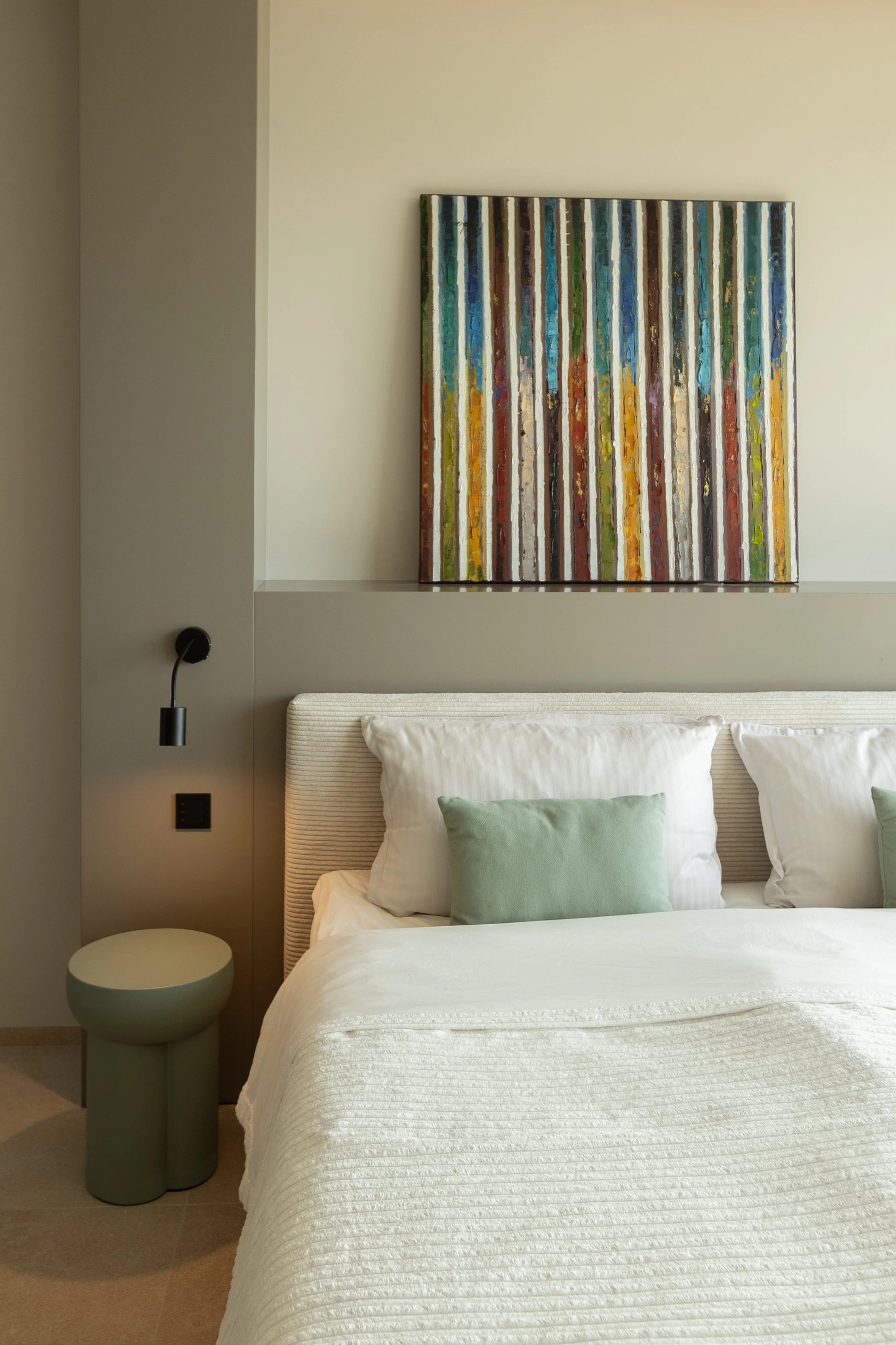 Habitación moderna con cama vestida con ropa blanca y dos almohadas, una de color verde y otra blanca, junto a una medianoche verde y una lámpara negra. Decoración minimalista con un cuadro abstracto en la pared y una pared en tono beige.
