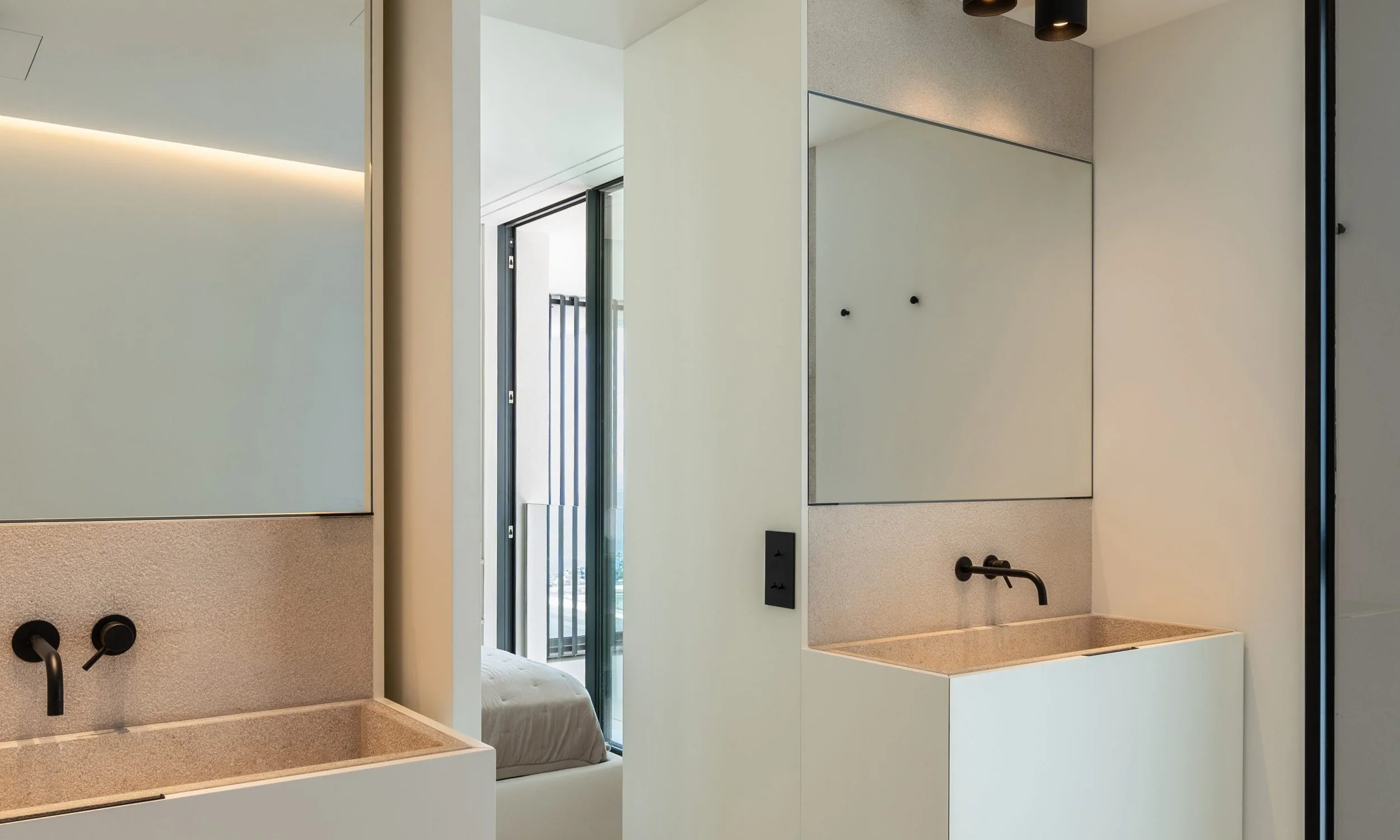 Espejo rectangular en una pared blanca, con un lavabo y grifo negro en primer plano, y una puerta de balcón o ventana corrediza que revela un balcón con barandilla y un cielo despejado.