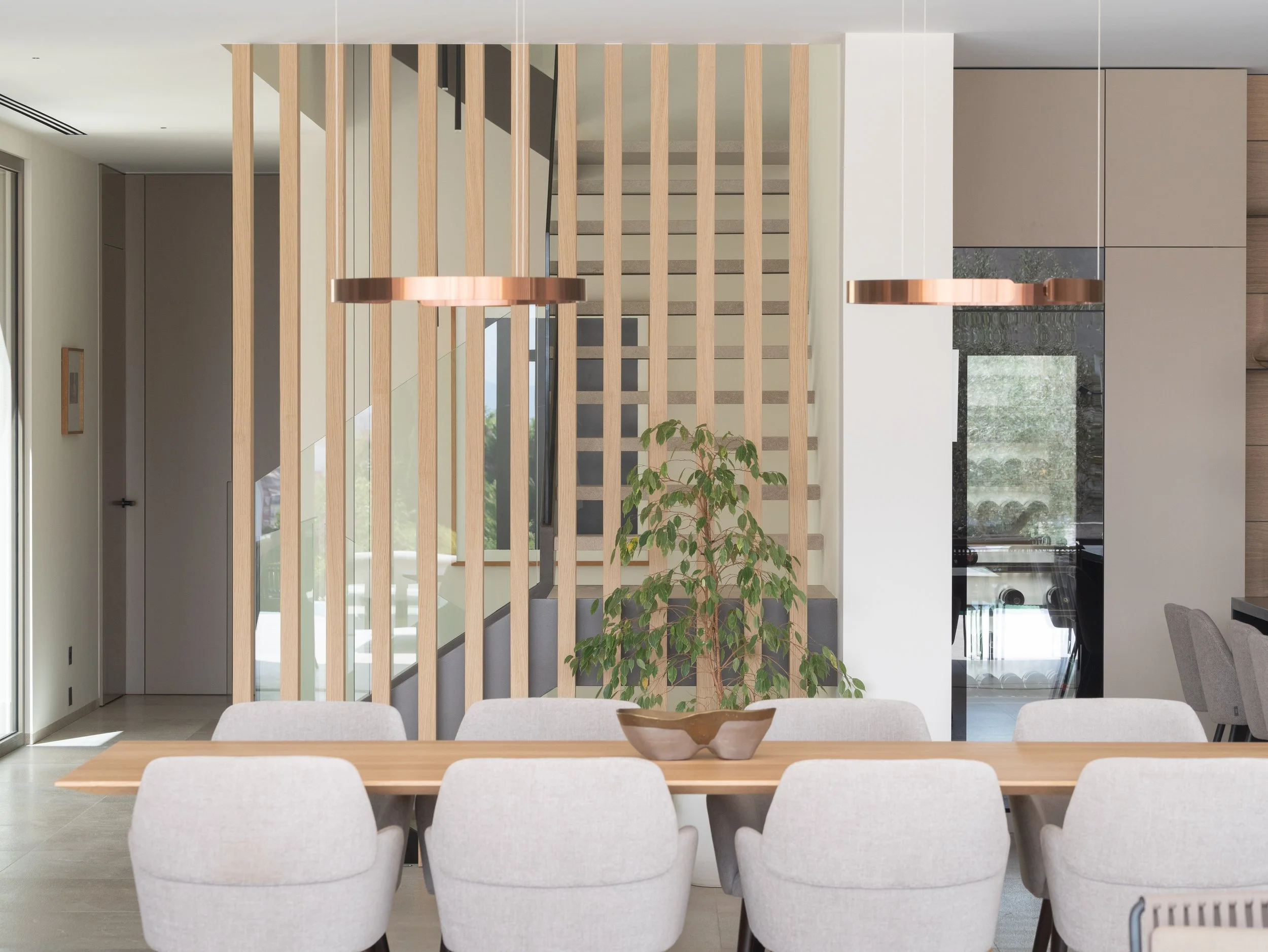 Vista interior de comedor moderno con mesa de madera y sillas blancas, división decorativa de listones de madera y plantas, zona de cocina en el fondo con gabinetes beige y encimera de piedra negra.