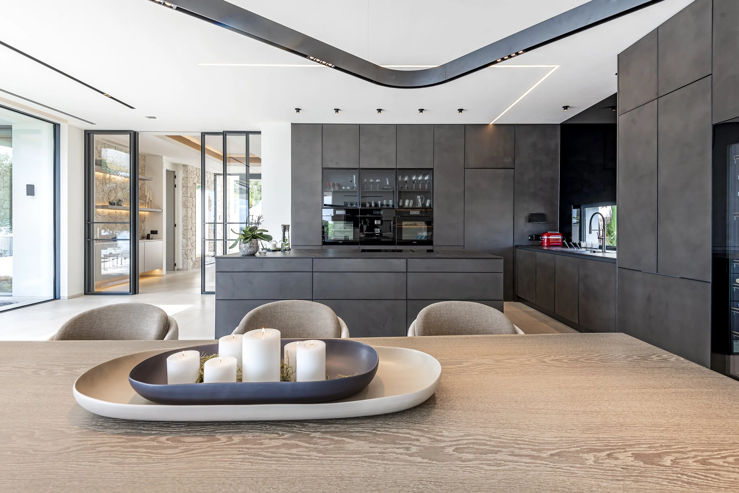 interior de una cocina moderna con muebles grises, una mesa de comedor con velas en un plato, y ventanas grandes que dejan entrar luz natural