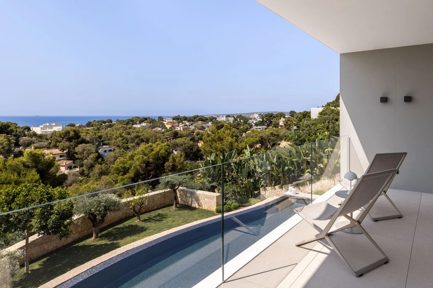 Vista desde un balcón moderno con sillas reclinables, disfrutando del paisaje de árboles, casas y el mar en el horizonte, en un día soleado.
