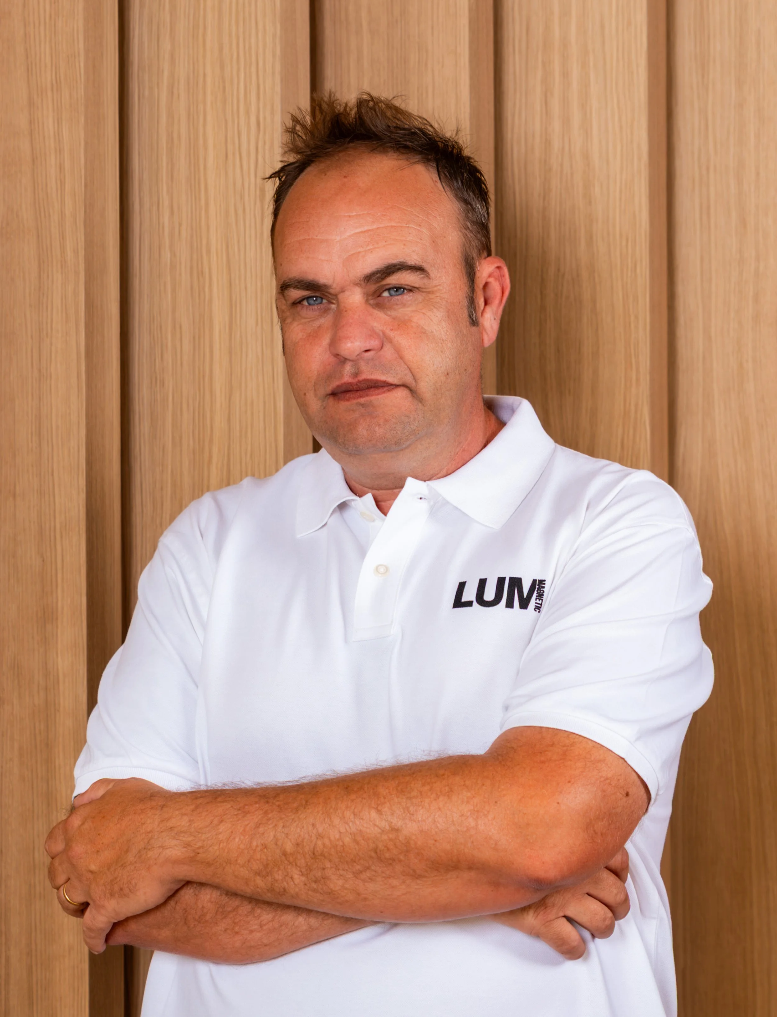 Hombre con camiseta blanca y fondo de paneles de madera, cruzado de brazos, con cabello corto y expresión confiada.