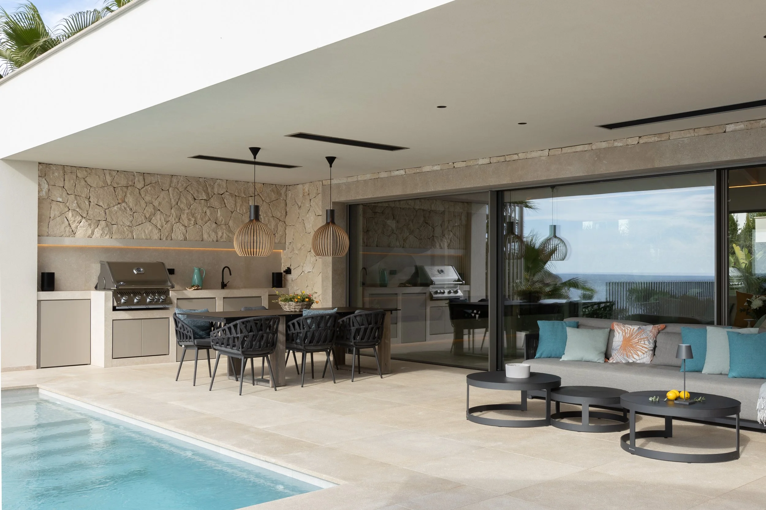Área de patio con piscina, una mesa con sillas, una sala con sofá y cojines, cocina exterior, y una vista del mar al fondo.