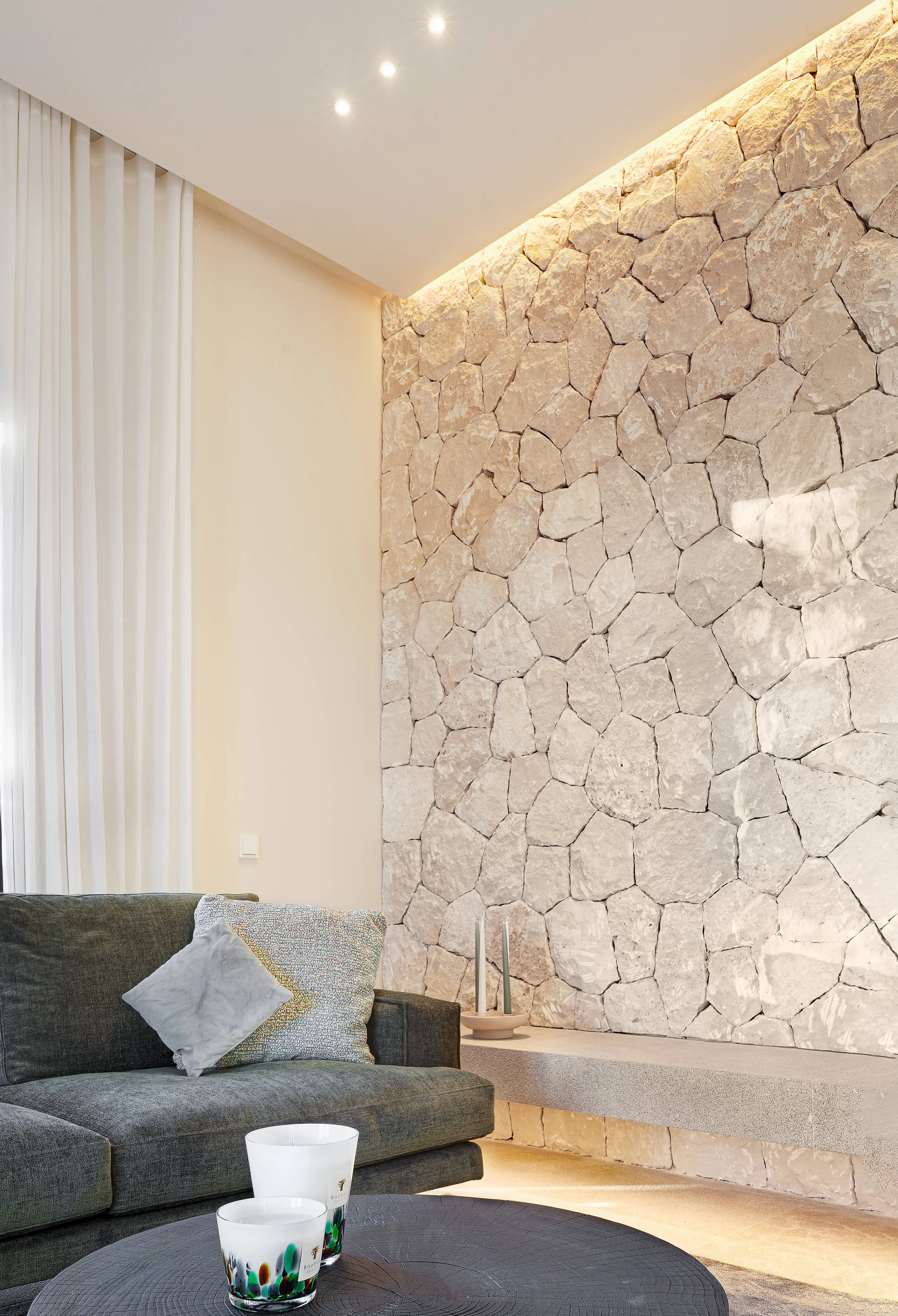 Sala moderna con pared de piedra natural, sofá gris con cojines, mesa de centro, y decoración minimalista.