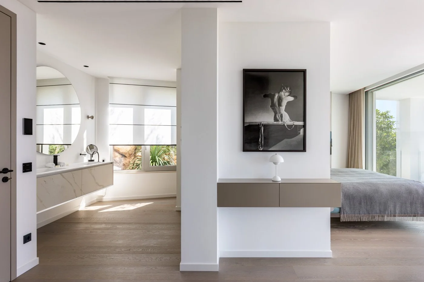 Vista interior de un apartamento moderno con dormitorio y baño, decorado en tonos neutros, con ventanas grandes y cortinas beige, y una obra de arte en la pared