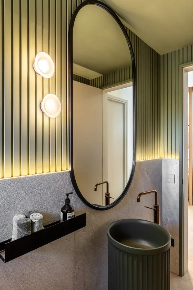 Consulta de baño con espejo ovalado grande, paredes con listones decorativos verdes y luz integrada, lavabo circular oscuro y grifo dorado. Espejo de pared, puerta de entrada y estantería con toallas y jabón.