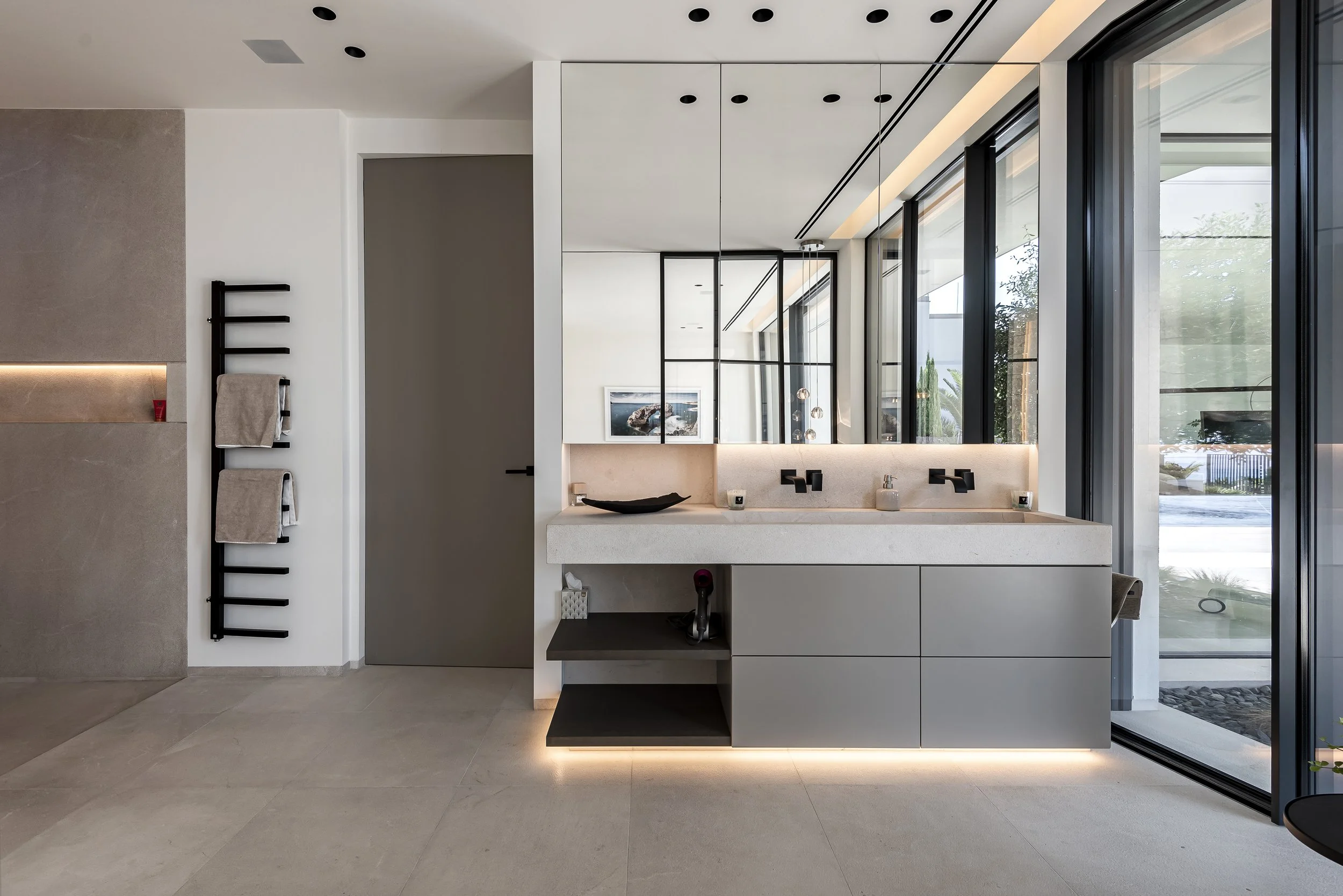 Interiores de baño moderno con lavabo de mármol, espejo grande con marcos de espejo, ventanas de piso a techo, iluminación ambiental, toallas colgadas en una estantería negra, y vista hacia un jardín exterior.