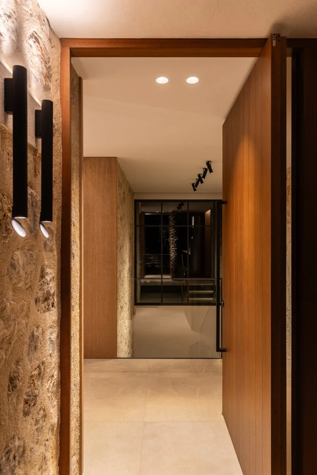 Vista de un pasillo interior con paredes de piedra y madera, iluminación empotrada en el techo y una puerta de cristal al fondo.