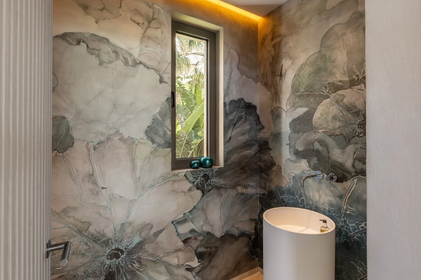 Baño con paredes decoradas con mármol de tonos gris y beige, ventana con vista a plantas verdes, lavabo de pedestal blanco y pequeños objetos decorativos sobre la ventana.