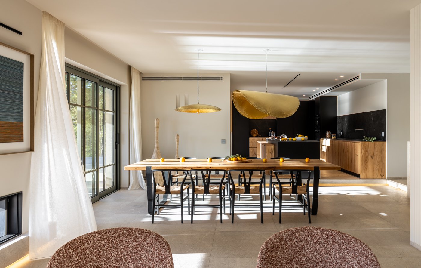 Sala de comedor moderna con mesa de madera, sillas negras, y decoraciones con naranjas y cortinas blancas, vista a la cocina con encimeras de madera y negro, grandes ventanas con cortinas blancas, luz natural.