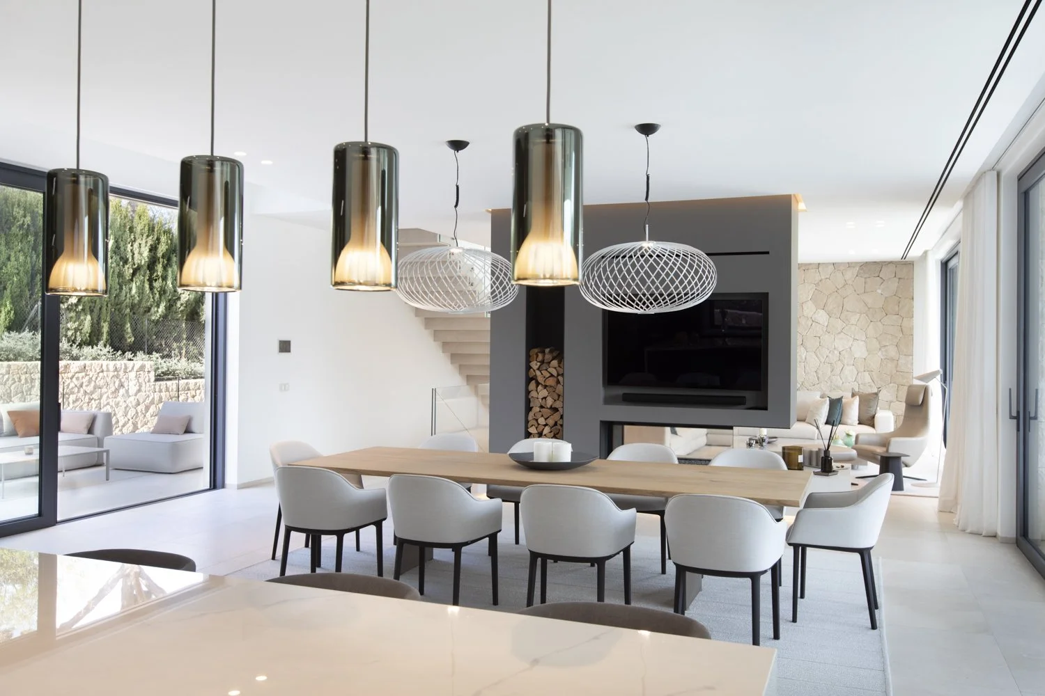 Interior de sala de comedor moderna con lámparas colgantes de diseño y muebles blancos, con salida a área exterior con sillas y mesa vista por ventanas grandes.
