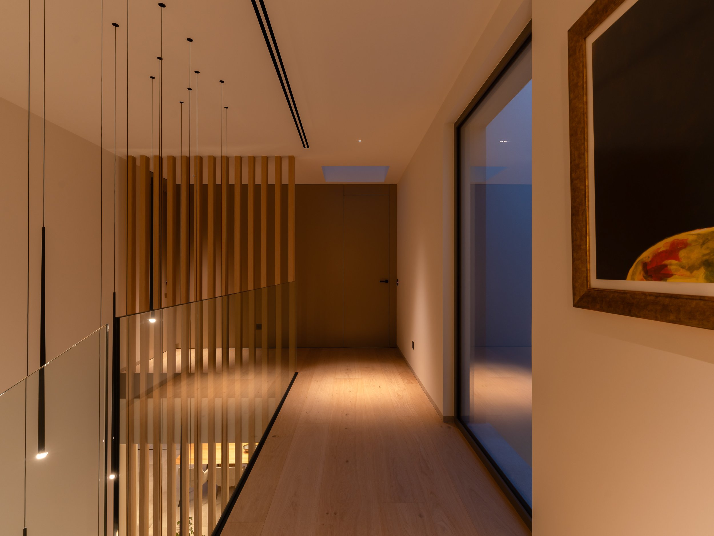 Vista interior moderna con paredes blancas, techos altos, piso de madera de color claro y elementos decorativos de madera. Fotos enmarcadas decoran la pared a la derecha y lámparas delgadas cuelgan del techo. La iluminación resalta el ambiente cálido