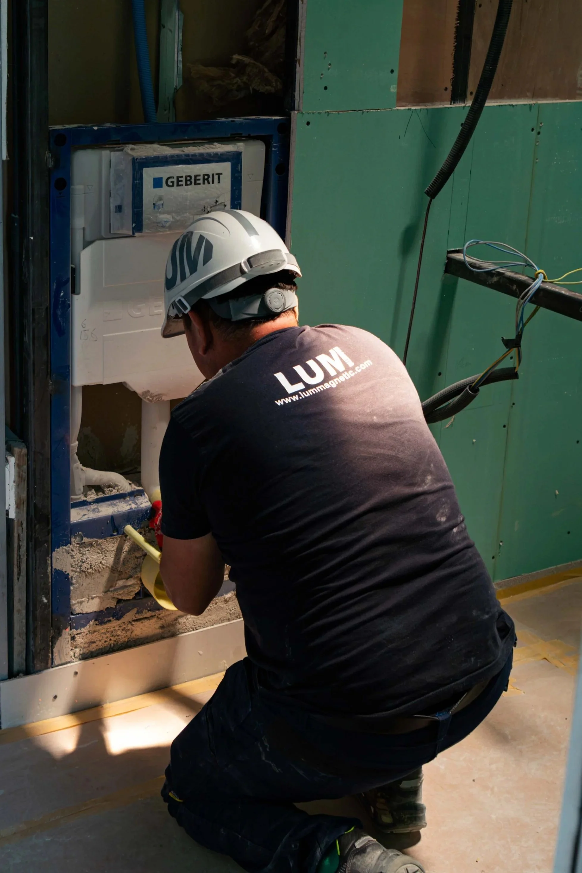 Técnico de construcción trabajando en la instalación de un inodoro en construcción, usando casco de seguridad y ropa de trabajo, en un espacio con pared de drywall verde y cables eléctricos visibles.