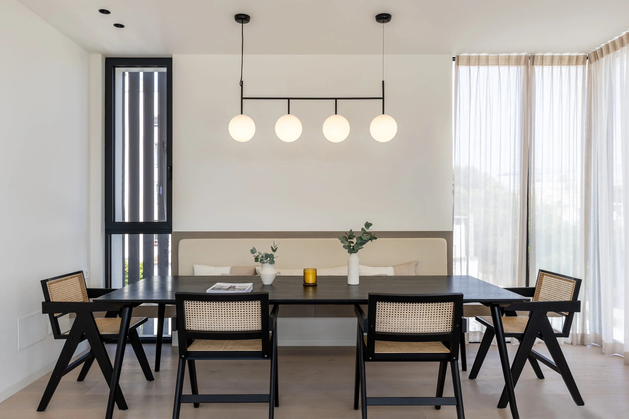 Mesa de comedor negra con cinco sillas de madera y ratán, con decoraciones de vasijas blancas, una amarilla y un libro. Fondo con cortinas blancas y ventanas. Lámpara de cuatro focos colgantes.