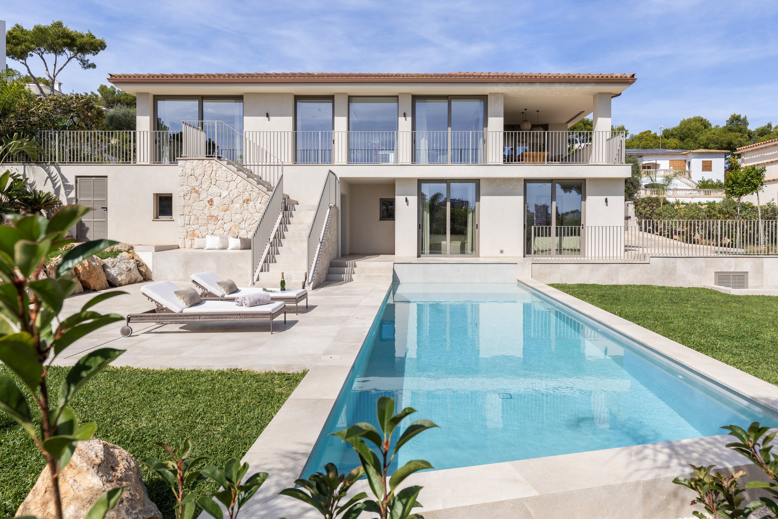Casa moderna con piscina, tumbonas y jardín verde, rodeada de árboles y con ventanas grandes.