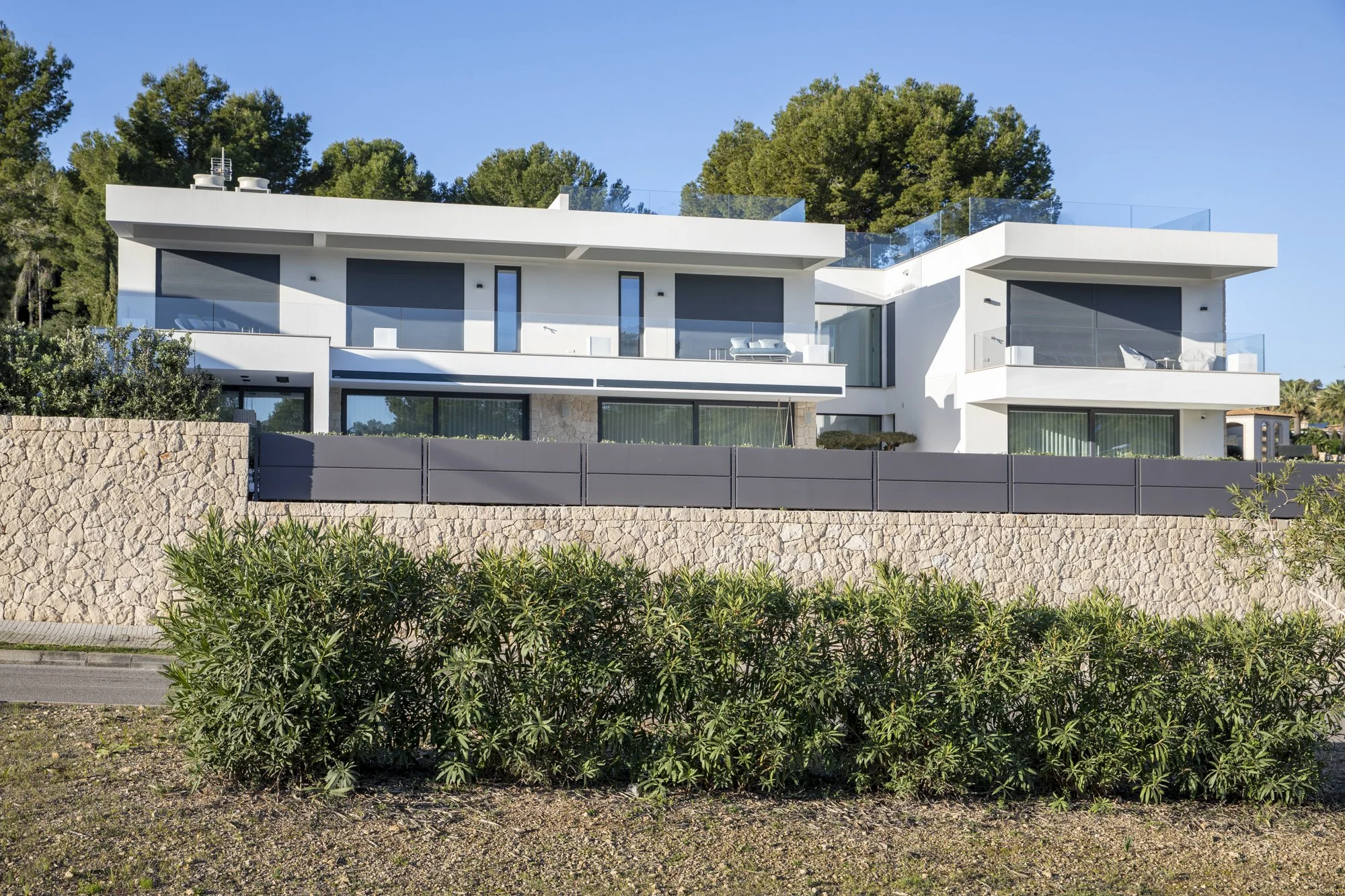 Casa moderna de varios pisos con grandes ventanas y balcones, rodeada de árboles y cercada por una pared de piedra y un cerco negro