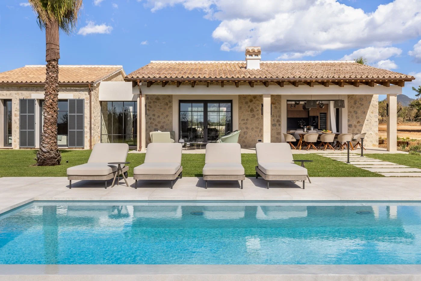 Casa de estilo rústico con piscina, tumbonas y área de comedor en el patio, en un entorno soleado y con cielo parcialmente nublado.