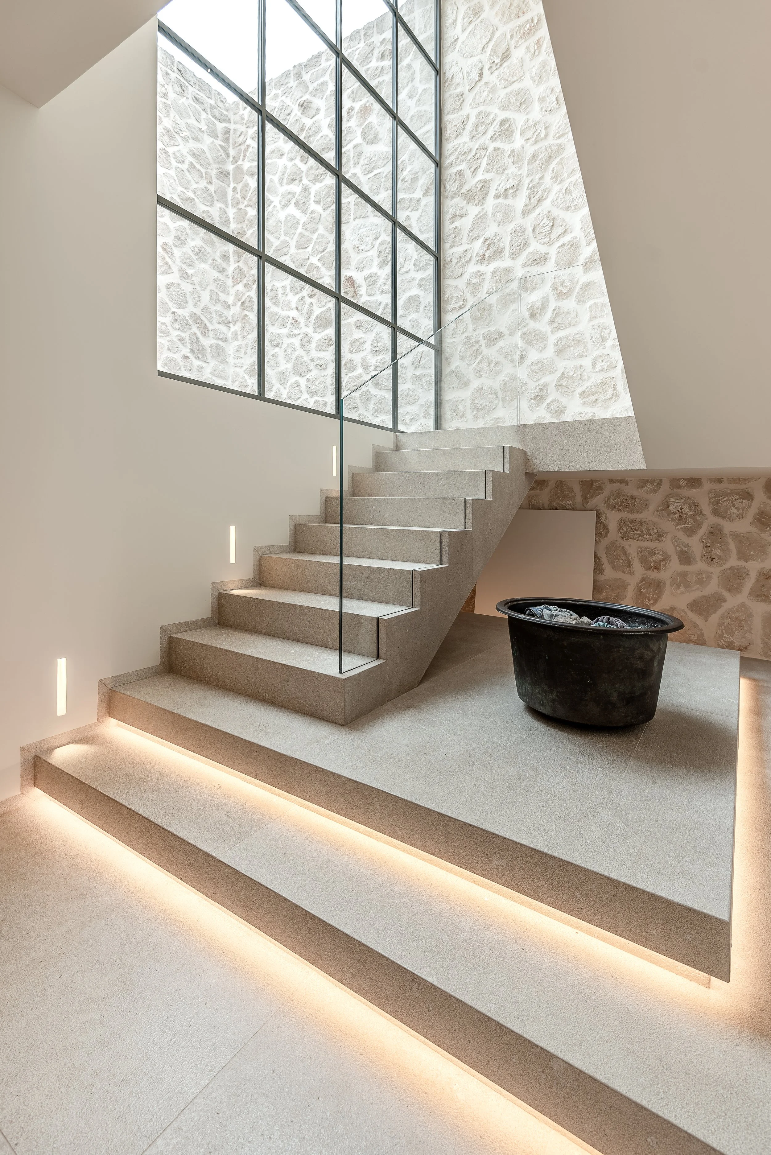 Escalera de concreto con iluminación empotrada, paredes de piedra blanca y gran ventanal de estructura metálica que permite la entrada de luz natural a un espacio moderno.