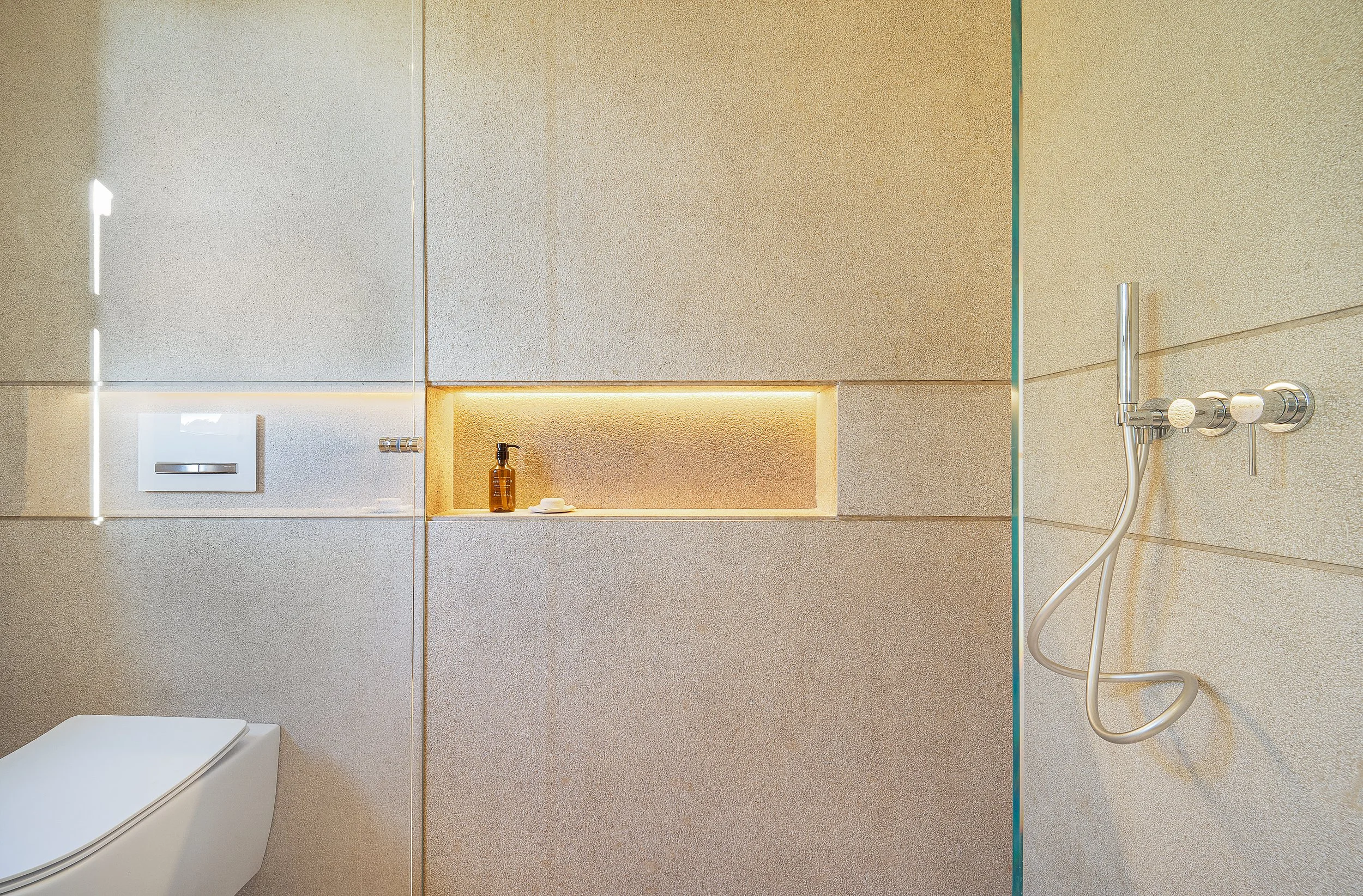Vista de una ducha moderna con paredes de azulejo beige, ducha de mano, dispensador de jabón y un inodoro en un baño minimalista.