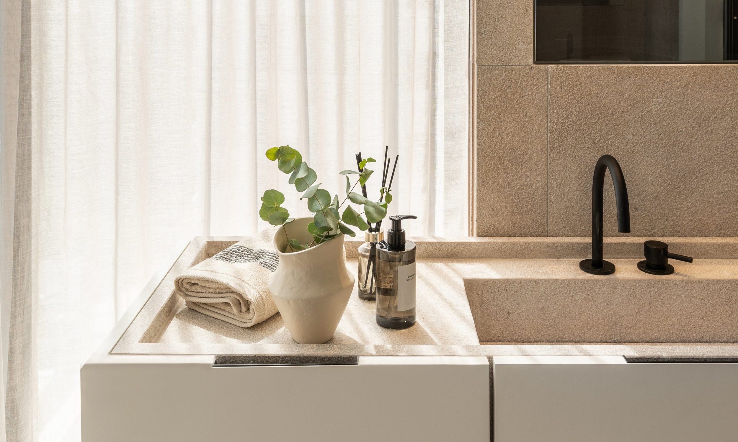 Lavabo de cocina con una planta en una jarra blanca, toallas blancas, y jabón en dispensador, frente a una ventana con cortinas blancas y una pared de azulejos beige.