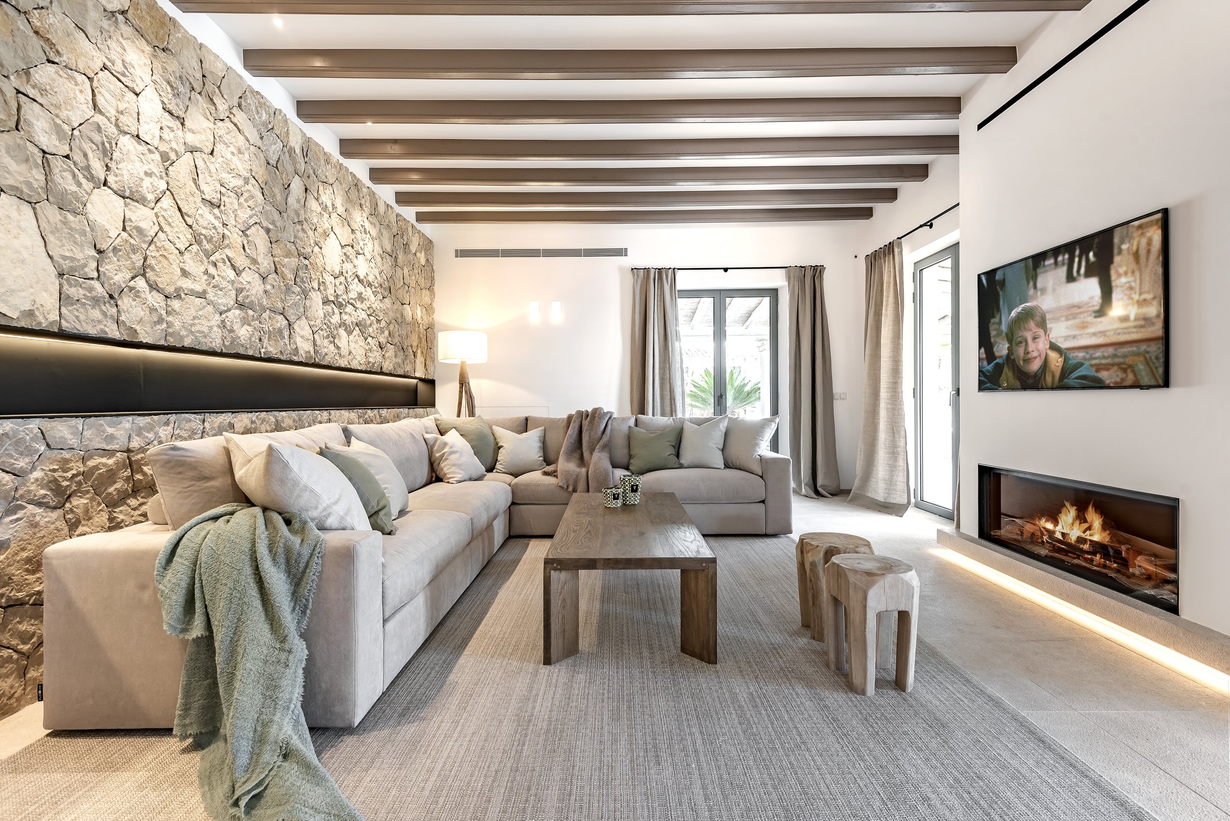 Sala moderna con sofá beige, chimenea de gas, televisor en la pared, paredes blancas, puerta de vidrio con cortinas y pared de piedra natural