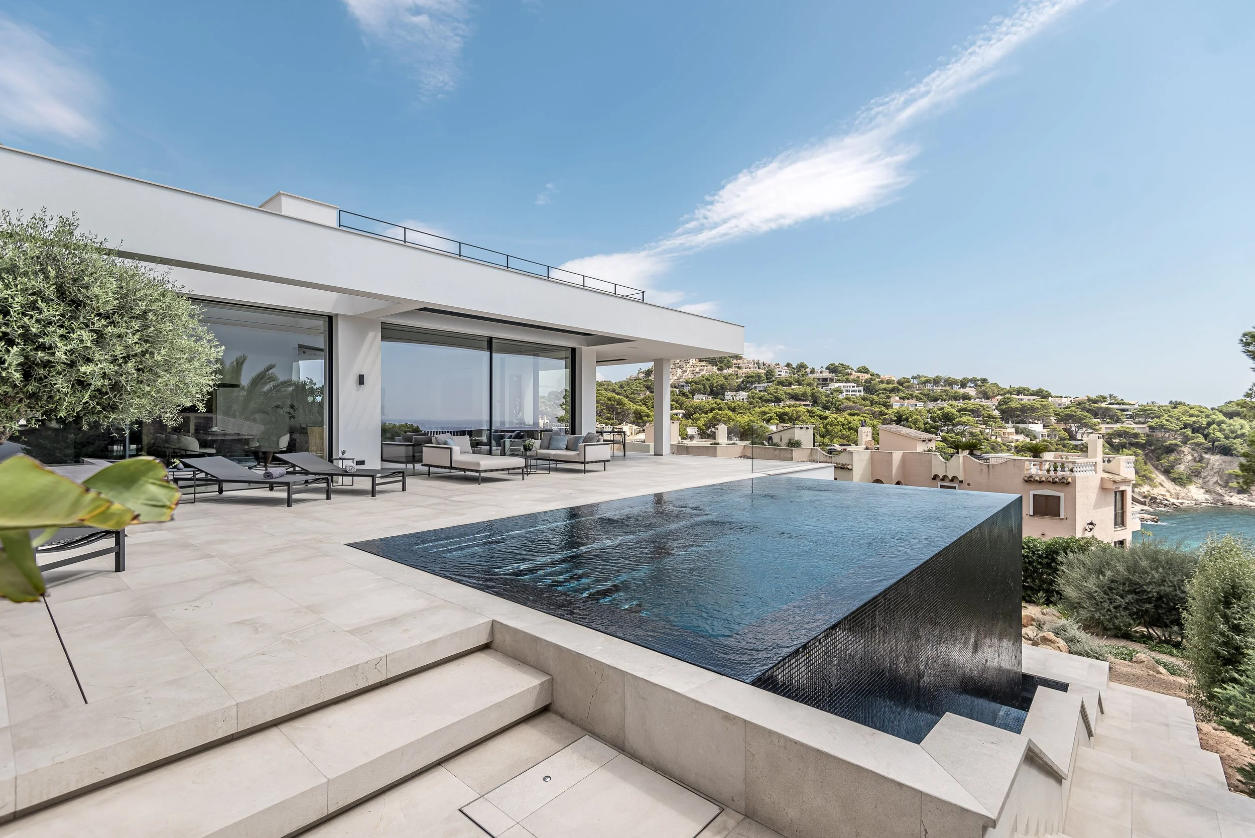 Vista de una casa moderna con terraza y piscina con vista al mar y colinas verdes en el fondo, cielo despejado y algunos edificios en las colinas.