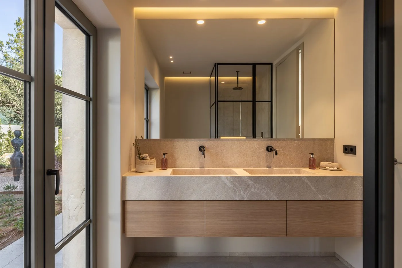 Lavabo doble con espejo grande, encimera de piedra y mueble de madera en baño moderno con ventanas grandes.