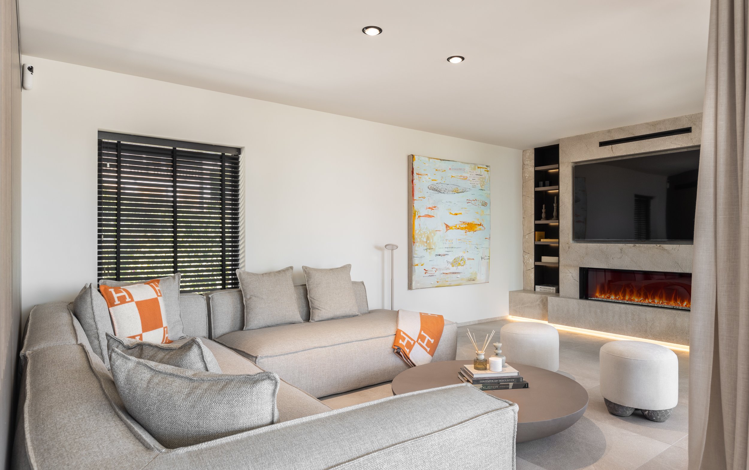 Sala moderna con sofá beige, cortinas negras, cuadro abstracto y chimenea eléctrica, con mesa de centro gris y decoraciones.