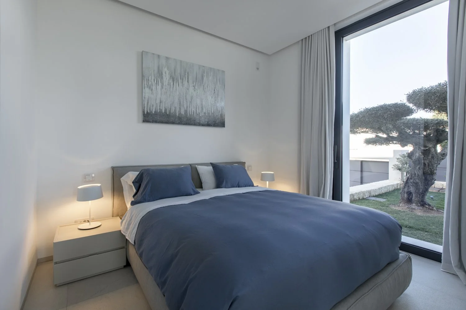 Habitación moderna con cama grande, ropa de cama azul, dos lámparas de noche y una vista de un árbol en un balcón con cortinas blancas