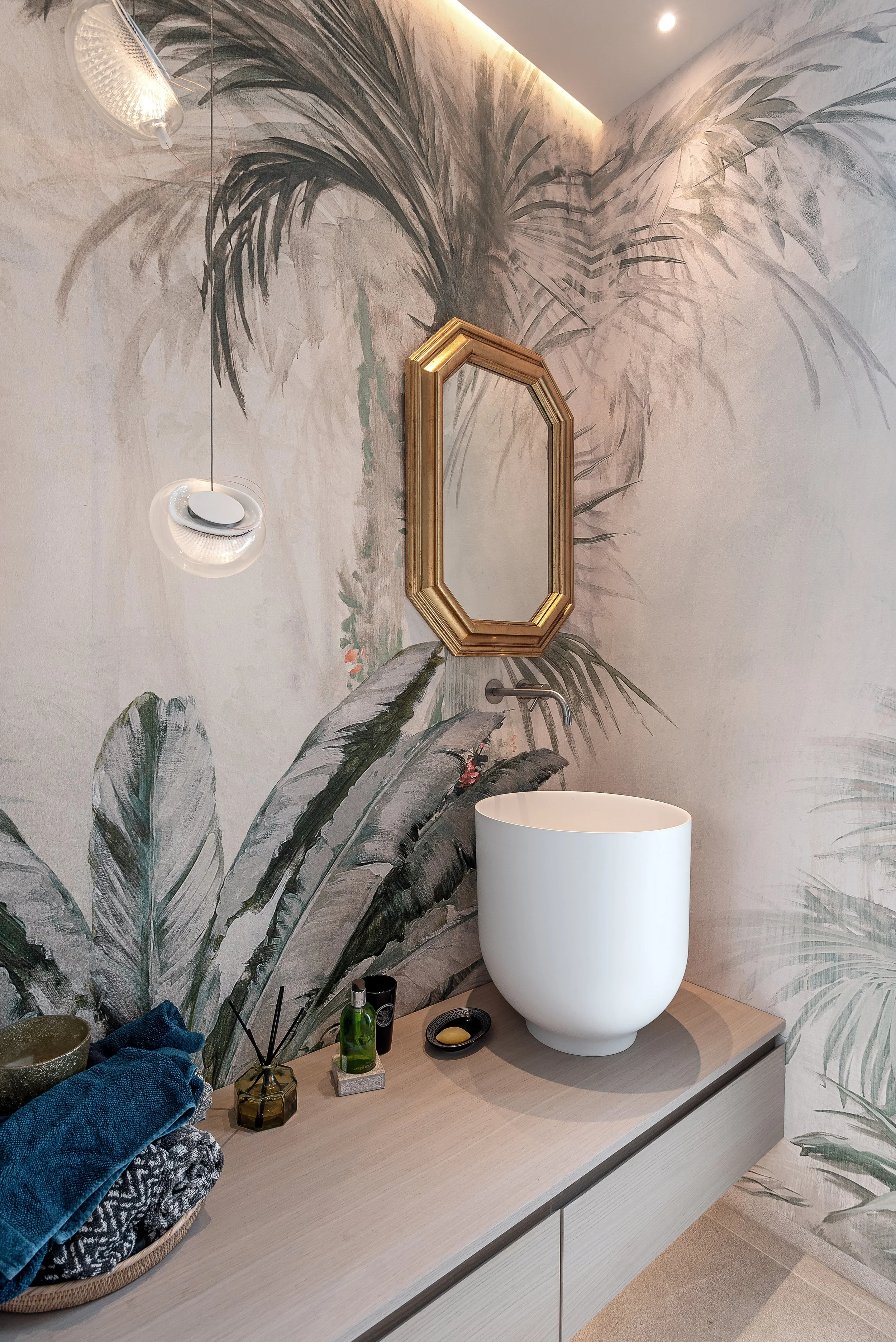 Espejo octogonal dorado en una pared decorada con mural de hojas en tonos verdes y grises, lavabo blanco de forma ovalada sobre mueble de madera clara, y accesorios de baño en la superficie.