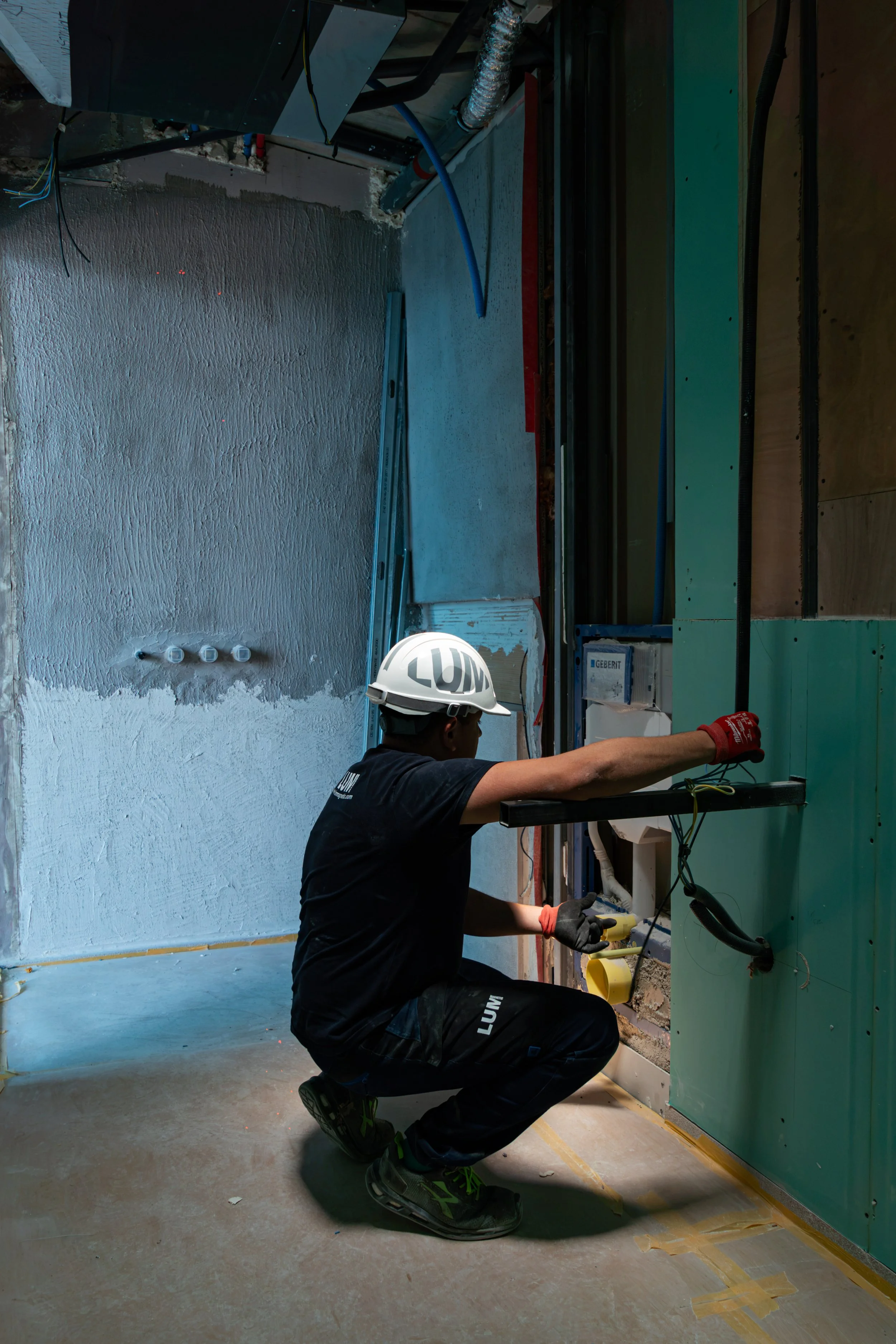 Trabajador instalando cableado eléctrico en una pared en construcción, llevado casco de seguridad y guantes, en un espacio interior con paredes de yeso y tuberías.