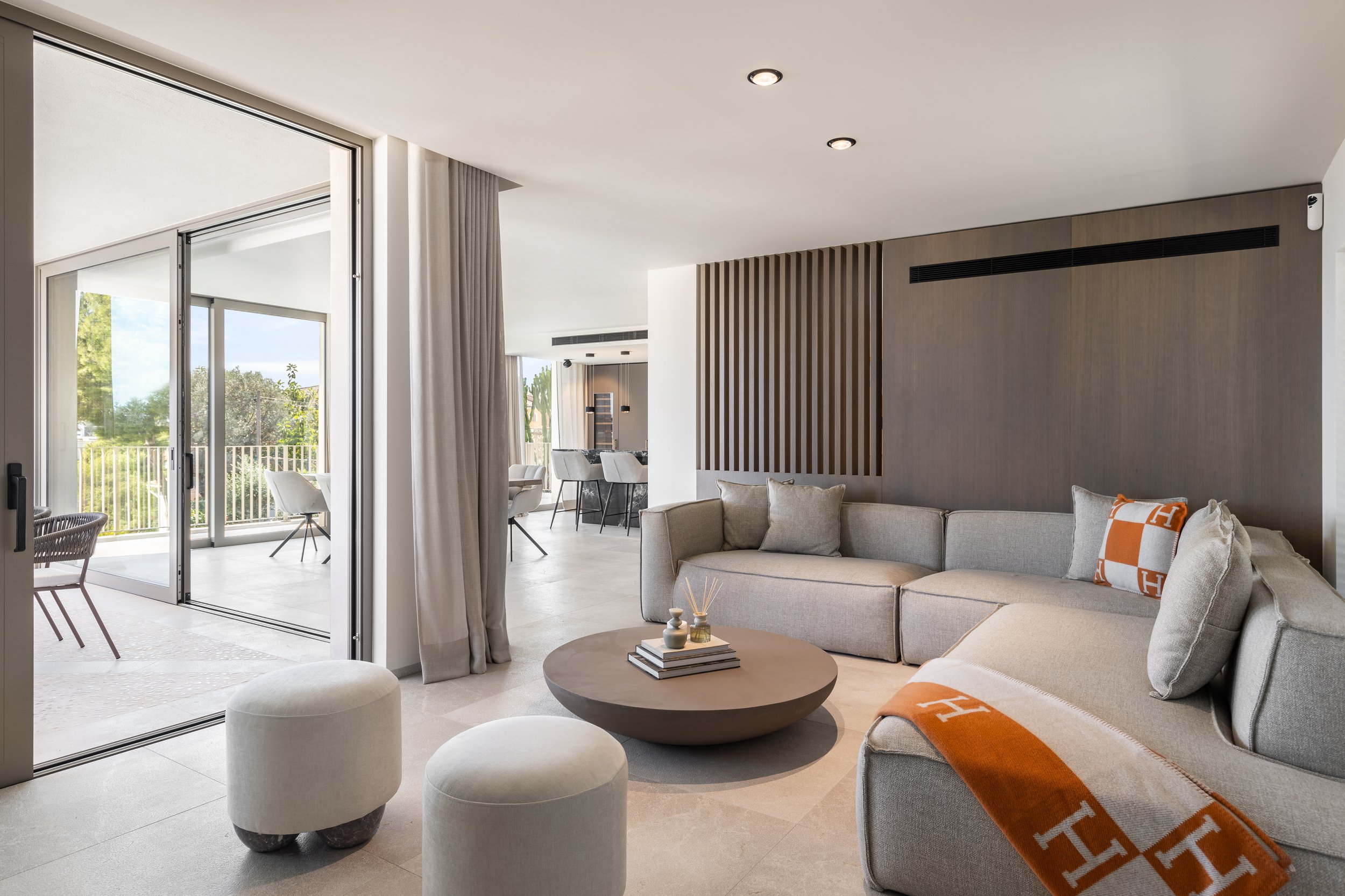 Sala de estar moderna con sofá beige, mesa de centro de madera, sillas en el comedor y puertas de vidrio que dan a un balcón con vista a árboles y cielo azul.