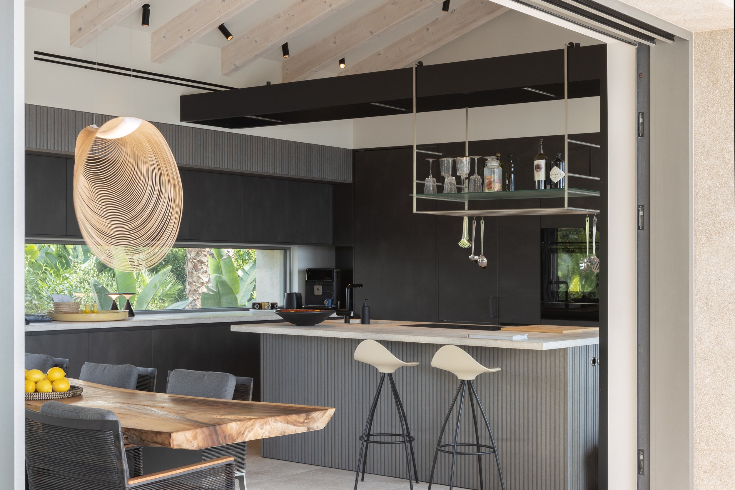 Cocina moderna con gabinetes negros, isla de madera con sillas, estantería de vidrio con botellas y vasos, ventana grande con vista a plantas tropicales, lámpara de diseño en forma de espiral de madera, techo de madera clara con luces empotradas.