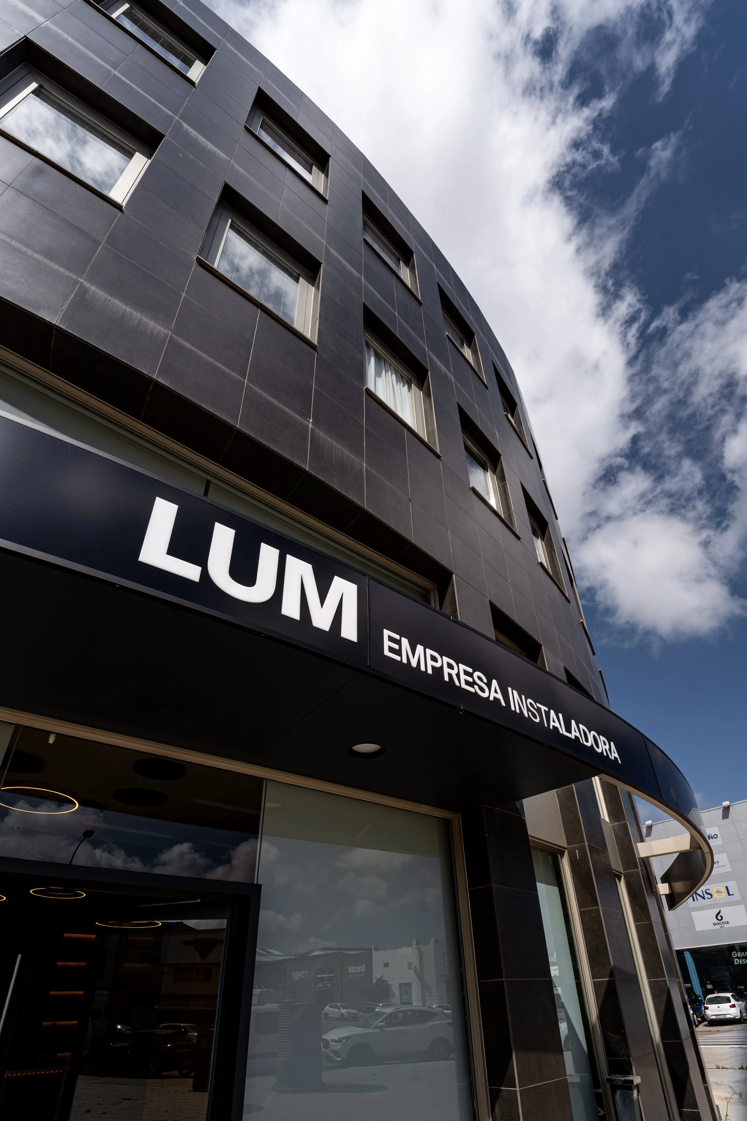 Vista del exterior de un edificio moderno de color negro con ventanas, bajo un cielo parcialmente nublado, con un cartel que dice 'LUM' y más texto que no se lee completamente.