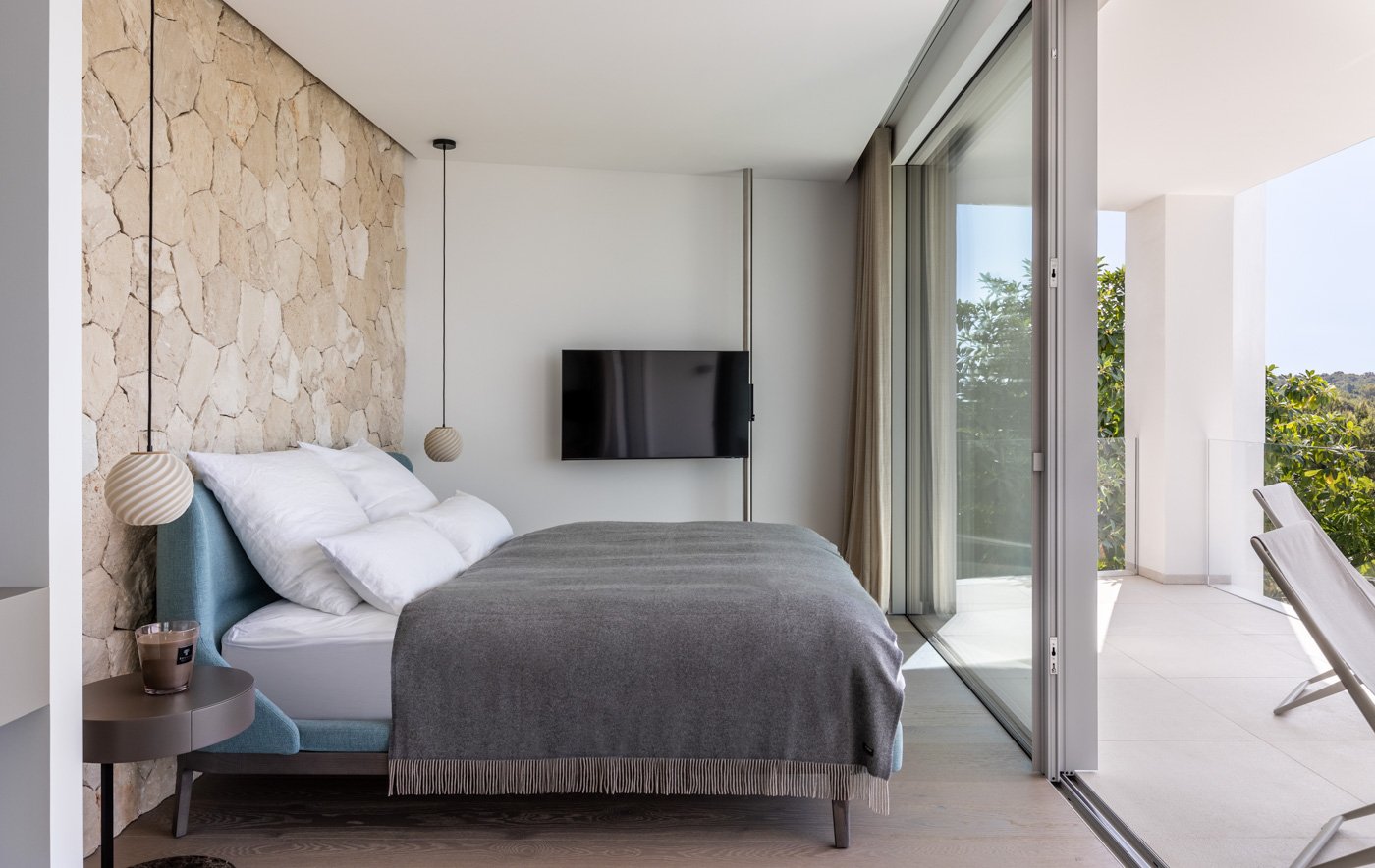 Habitación moderna con cama, paredes de piedra y vista a un balcón con sillas y árbol afuera.