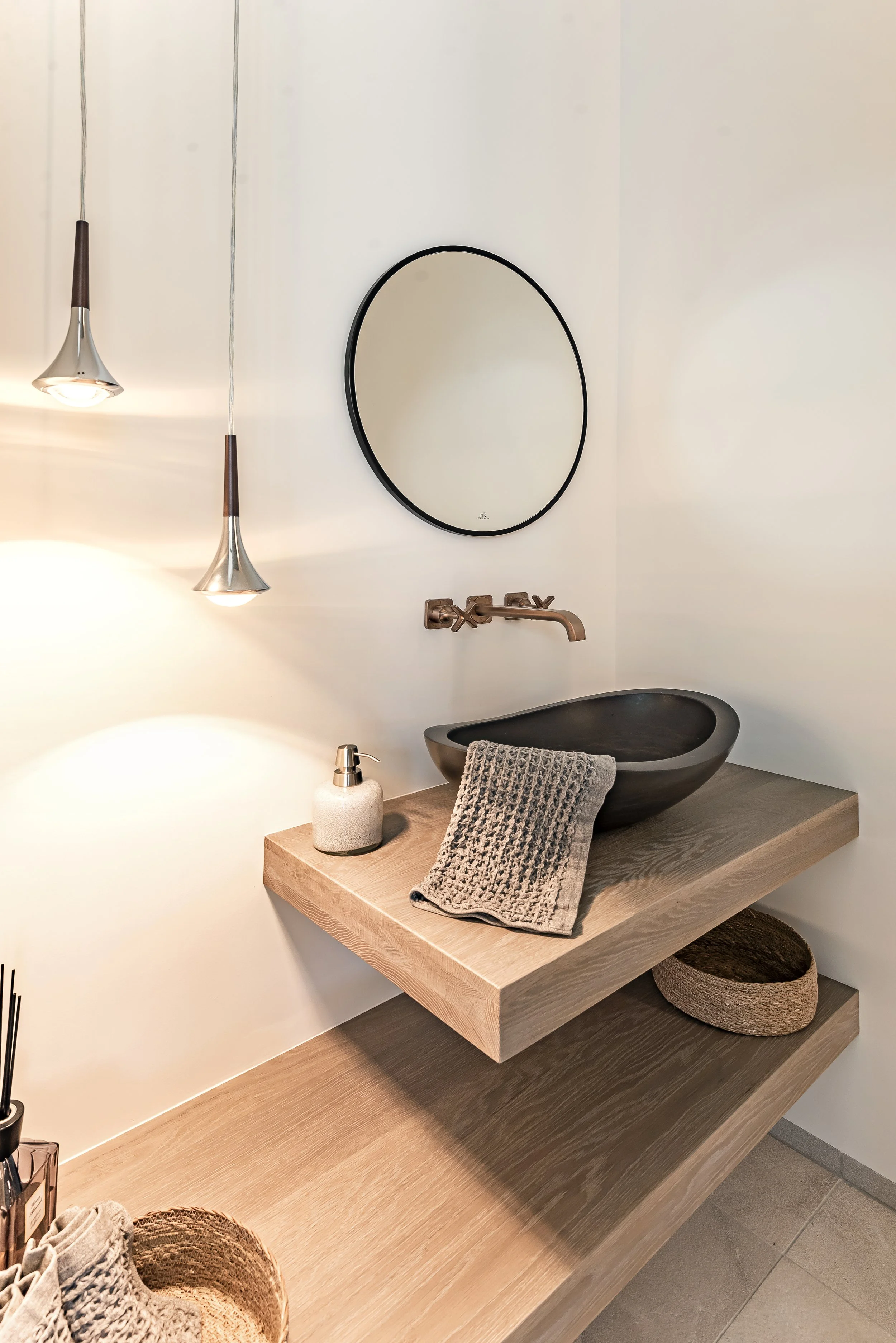 Espejo ovalado, lámparas colgantes, lavabo negro de piedra, grifería moderna, toalla gris sobre encimera de madera, accesorios de baño, cesta de piedra con toallas, ambiente de baño moderno y minimalista.
